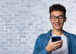 Smiling black curly hair funny young man, guy, cute boy wear braces hold hand typing text, looking reading gazing at cell phone mobile smartphone cellphone, stand against white bricks loft wall background