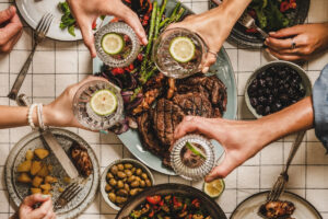 Summer barbeque party. Flat-lay of table with grilled meat, vegetables, salad, roasted potato and peoples hands feasting over checkered tablecloth, top view.